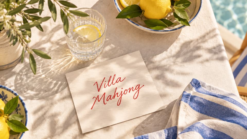 A sunny poolside table with bowls of lemons, lemonade, lemon blossoms, a blue-and-white striped napkin, and a handwritten Villa Mahjong card
